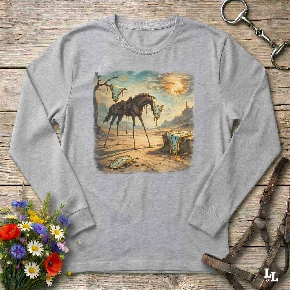 A light gray long-sleeve t-shirt with a square graphic print of a sad, broken horse in a desert landscape. The shirt is displayed flat on a rustic wooden surface, styled with equestrian leather tack, a bouquet of colorful wildflowers, and a metal horse bit.