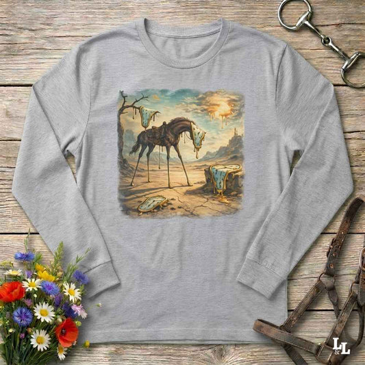 A light gray long-sleeve t-shirt with a square graphic print of a sad, broken horse in a desert landscape. The shirt is displayed flat on a rustic wooden surface, styled with equestrian leather tack, a bouquet of colorful wildflowers, and a metal horse bit.