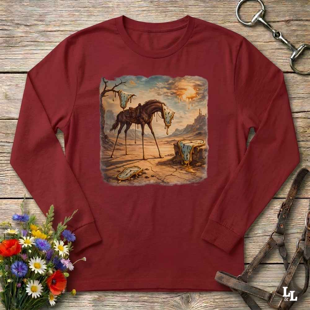 A long-sleeve crewneck t-shirt in maroon red, laid flat on a rustic wooden surface. The center of the shirt features a square graphic print depicting a surreal, vintage-style scene of a horse standing in a desert landscape with a sunset.