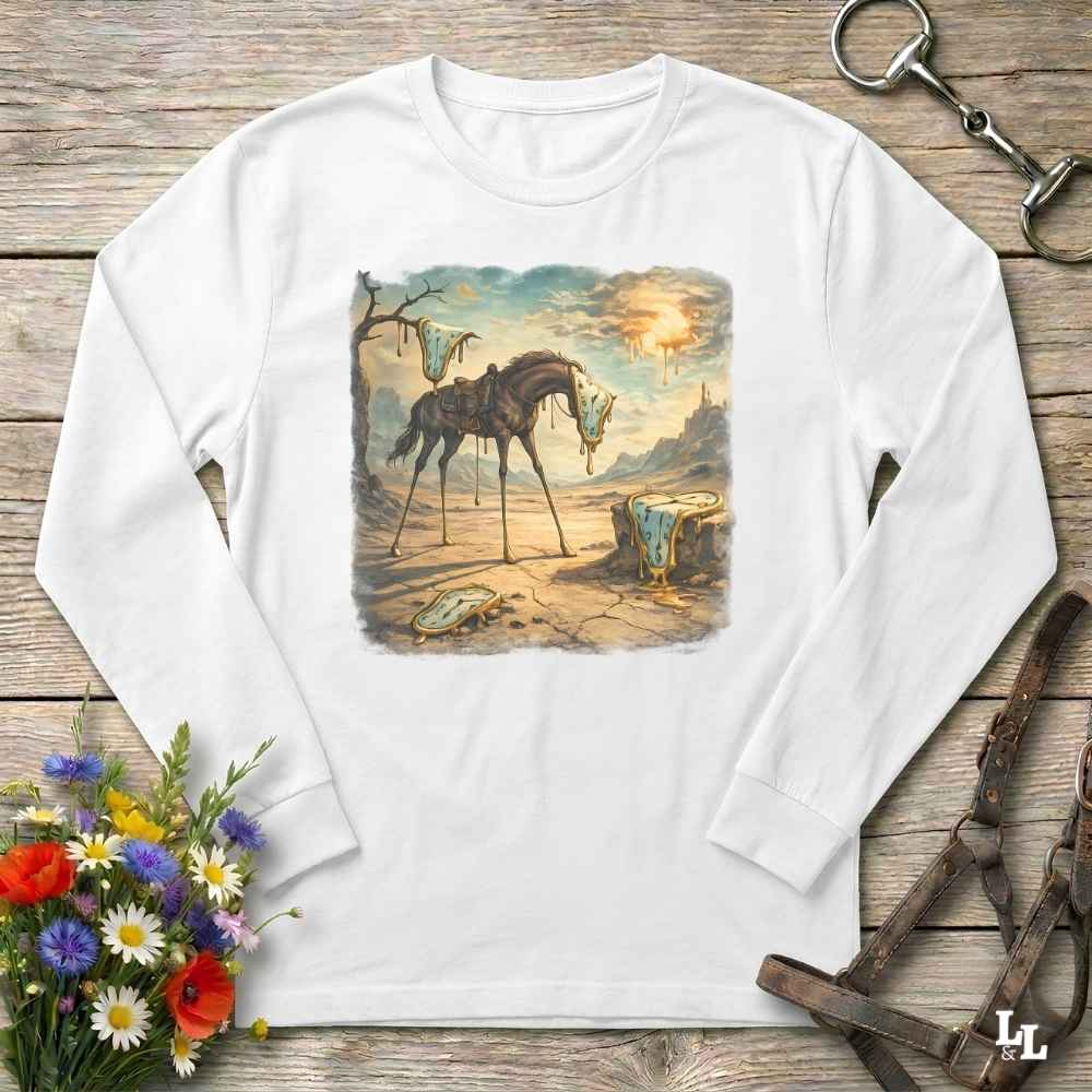 A white long-sleeve t-shirt with a vintage-style graphic print of a skeletal horse in a desert landscape, displayed flat on a rustic wooden background with equestrian accessories and flowers.