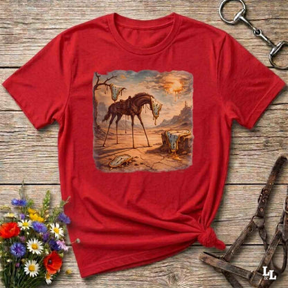A red short-sleeve t-shirt with a square graphic print of a cowboy riding a horse through a desert landscape, styled on a rustic wooden background with flowers and leather tack.