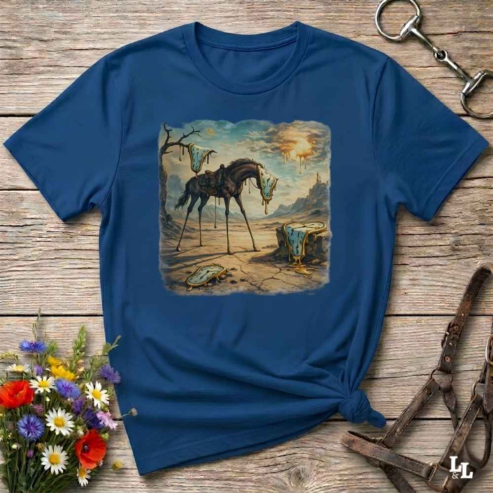 A navy blue short-sleeve t-shirt with a square graphic print of a surreal painting showing a horse standing in a desert landscape. The t-shirt is styled in a flat-lay on a rustic wooden background with equestrian leather tack and a small bouquet of colorful wildflowers.