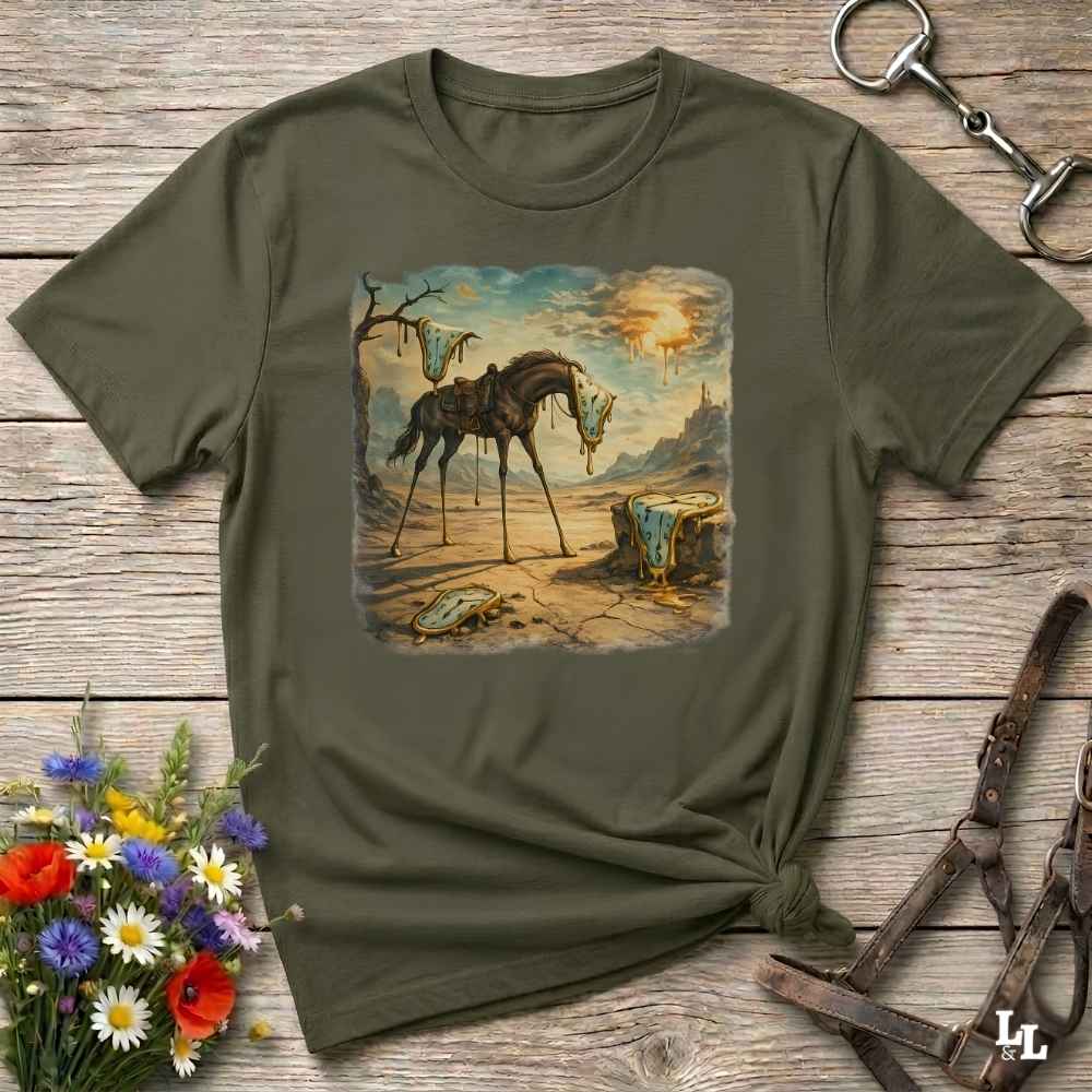 An olive green short-sleeve t-shirt with a square graphic print of a skeletal horse in a desert landscape, styled on a rustic wooden background with equestrian accessories and flowers.