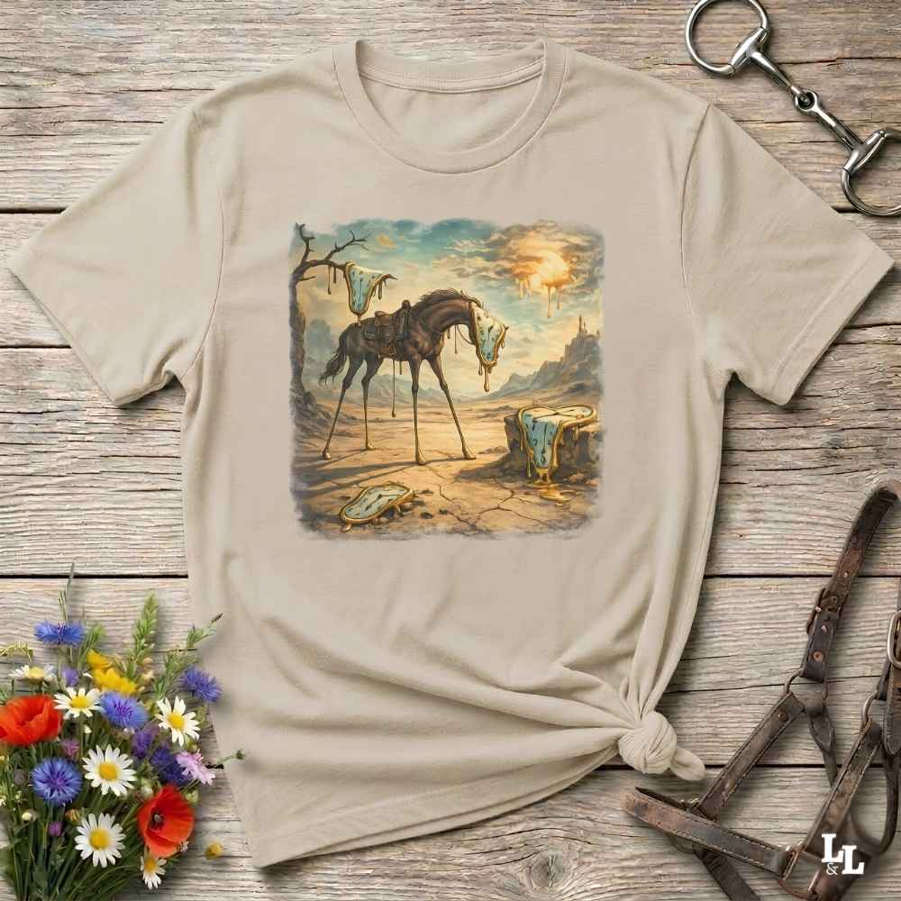 A beige graphic t-shirt with a vintage-style illustration of a horse in a desert landscape, styled in a flat-lay on a rustic wooden background with equestrian accessories and flowers.