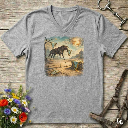 A heather gray V-neck t-shirt laid flat on a rustic wooden surface, featuring a square graphic print of a surreal desert landscape with a horse and a glowing sun. The shirt is styled with equestrian accessories (a horse bit in the top right and a halter in the bottom right) and a small bouquet of colorful wildflowers in the bottom left.