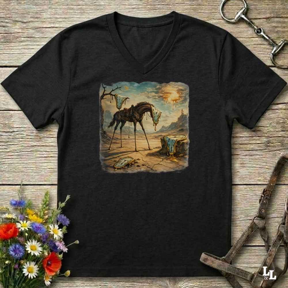 A black V-neck t-shirt laid flat on a rustic wooden surface, featuring a square graphic print of a surreal, dreamlike desert scene with a horse and a small figure. The shirt is styled with equestrian tack, a horse bit, and a bouquet of wildflowers.