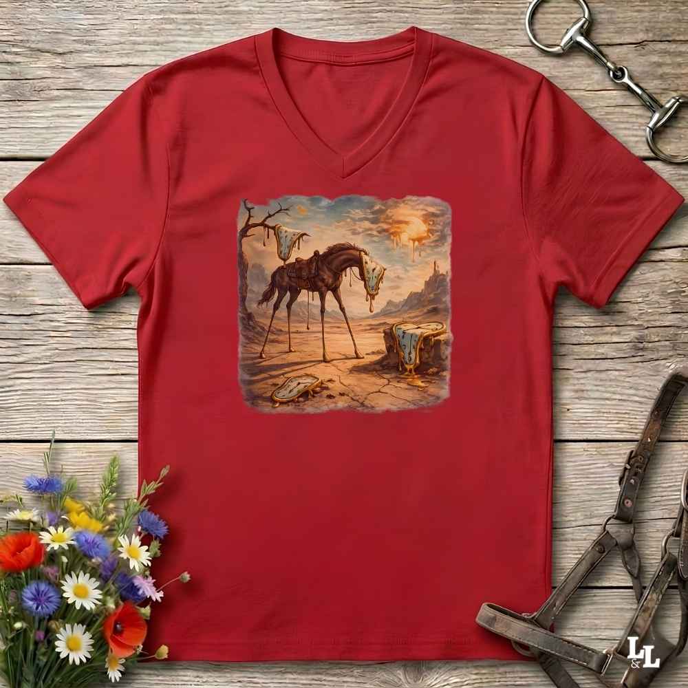 A red V-neck t-shirt laid flat on a rustic wooden surface, featuring a square graphic print of a surreal, dreamlike desert scene with a horse and a rider. The shirt is styled with equestrian accessories (a horse bit and reins) and a small bouquet of colorful wildflowers.