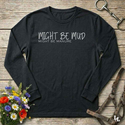 A black long-sleeve t-shirt with the text 'MIGHT BE MUD' and 'MIGHT BE MANURE.' printed in white on the chest, laid flat on a rustic wooden surface with equestrian and floral props.