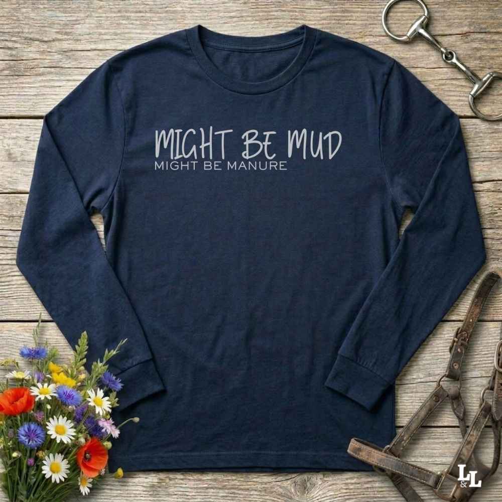A navy blue long-sleeve t-shirt with white printed text reading 'MIGHT BE MUD' and 'MIGHT BE MANURE' on the chest, laid flat on a rustic wooden background with decorative flowers and horse tack around it.