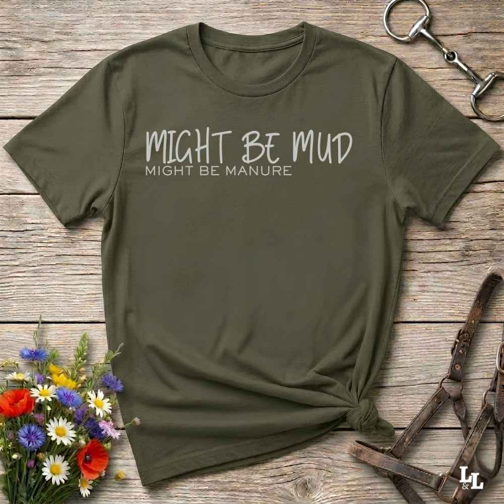 An olive green short-sleeve t-shirt with the text 'MIGHT BE MUD' and 'MIGHT BE MANURE' printed in white on the chest, styled in a flat-lay on a rustic wooden background with equestrian accessories and flowers.
