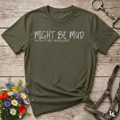 An olive green short-sleeve t-shirt with the text 'MIGHT BE MUD' and 'MIGHT BE MANURE' printed in white on the chest, styled in a flat-lay on a rustic wooden background with equestrian accessories and flowers.