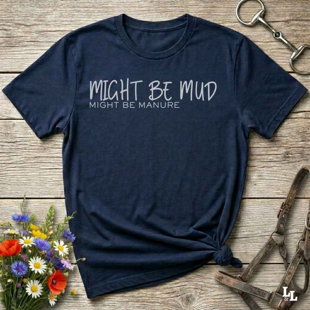 A navy blue  short-sleeve t-shirt with the text 'MIGHT BE MUD' and 'MIGHT BE MANURE' printed in white on the chest, styled flat on a rustic wooden background with equestrian accessories and flowers.