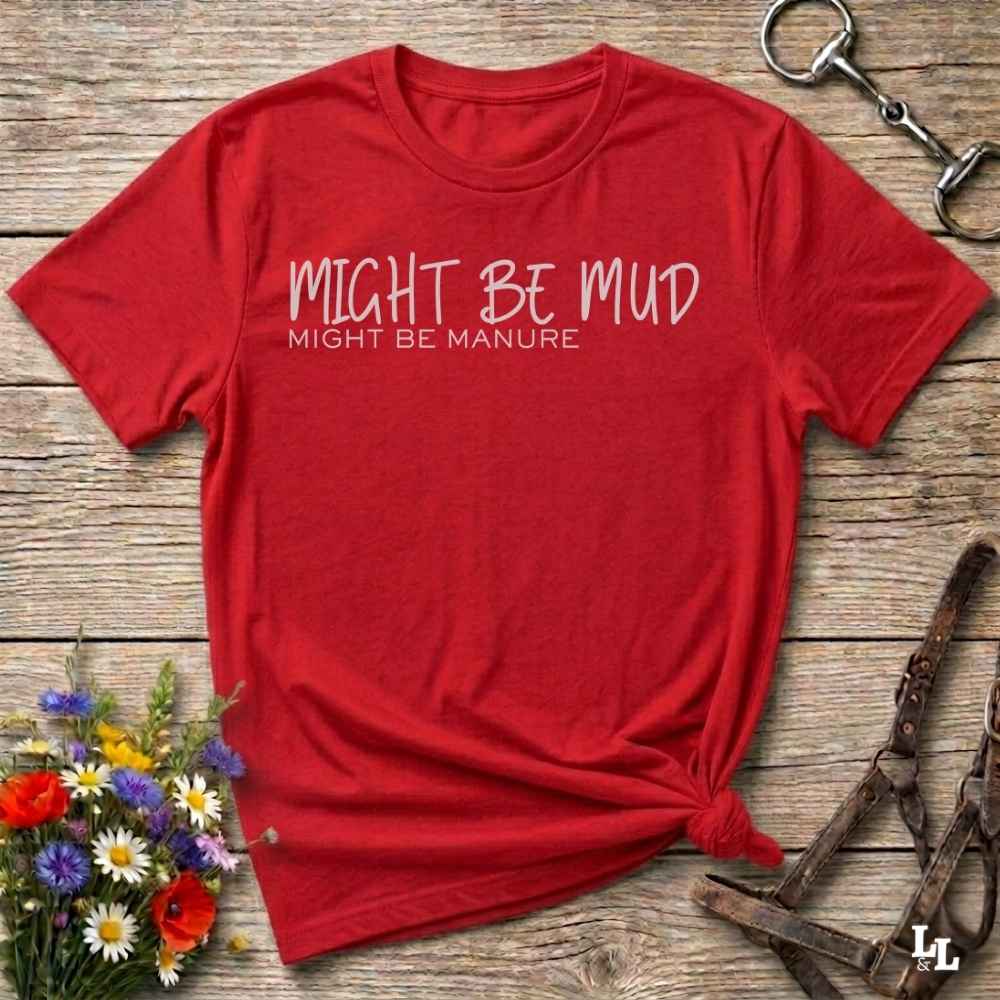 A red short-sleeve t-shirt with the text 'MIGHT BE MUD' and 'MIGHT BE MANURE' printed in white on the chest, styled flat on a rustic wooden background with equestrian accessories and flowers.