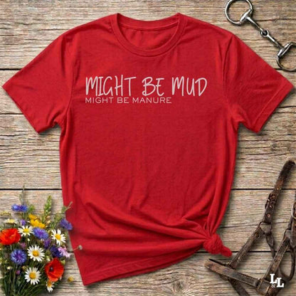 A red short-sleeve t-shirt with the text 'MIGHT BE MUD' and 'MIGHT BE MANURE' printed in white on the chest, styled flat on a rustic wooden background with equestrian accessories and flowers.