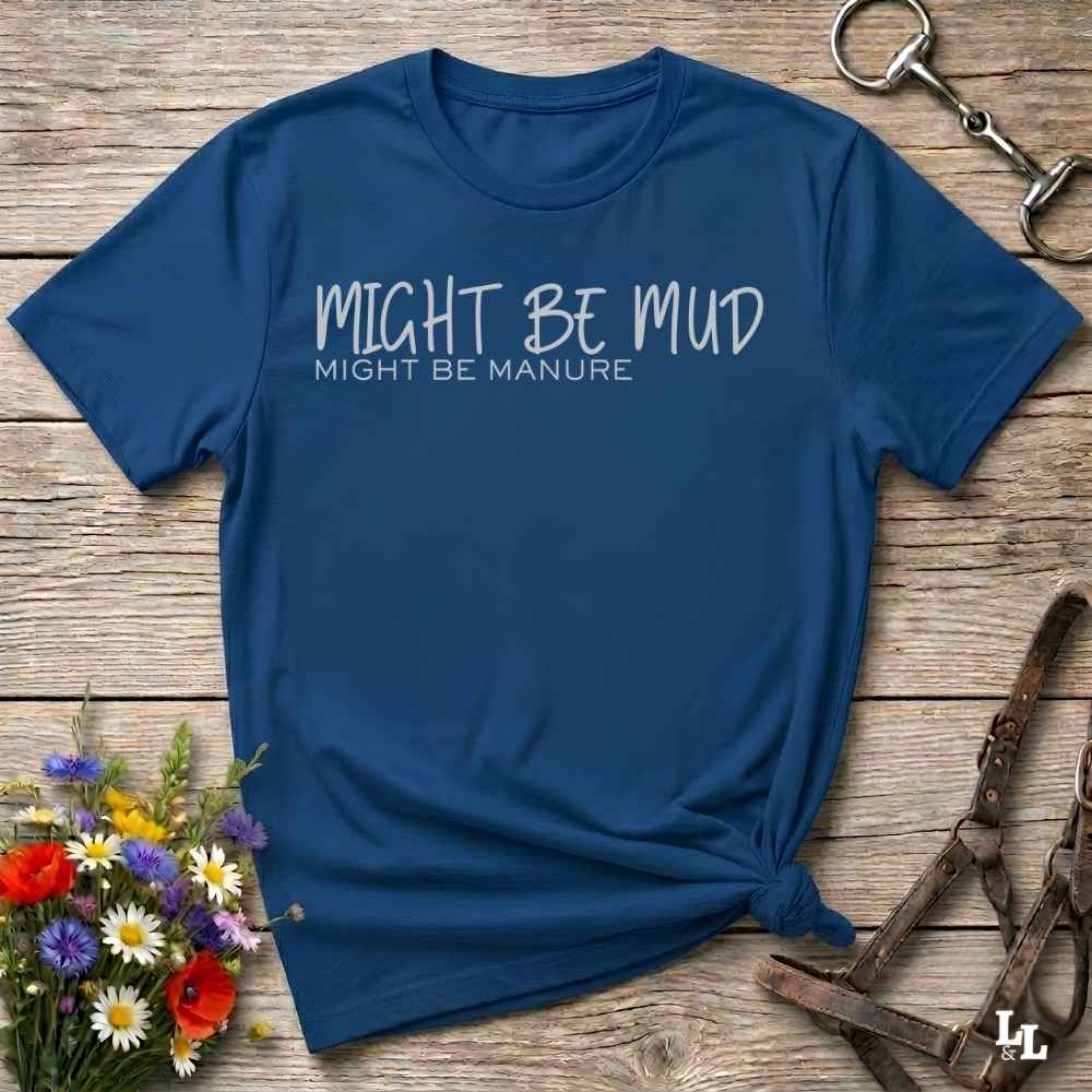 A navy blue short-sleeve t-shirt with the text 'MIGHT BE MUD' and 'MIGHT BE MANURE' printed in white on the chest, styled flat on a wooden surface with flowers, a horse bit, and a leather halter.