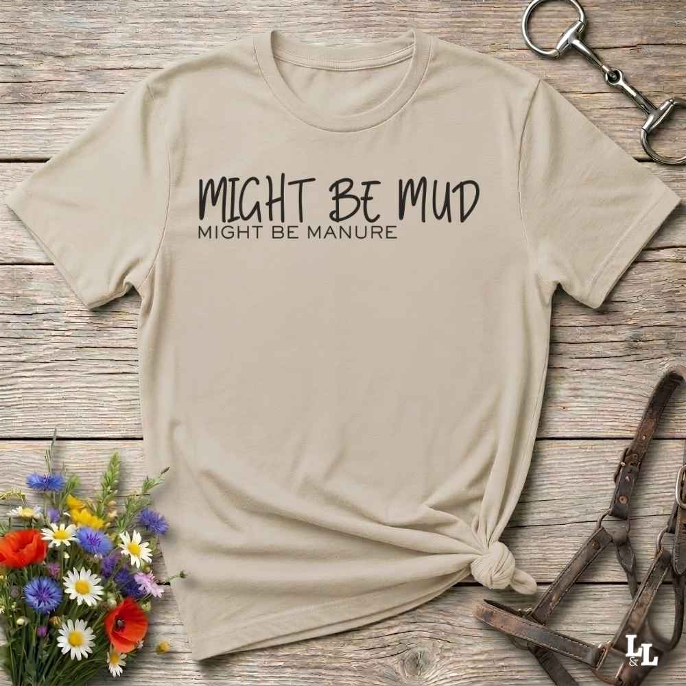 A beige graphic t-shirt with the text 'MIGHT BE MUD / MIGHT BE MANURE' printed in black, styled in a flat-lay on a rustic wooden background with a horse bit, horse halter, and wildflowers.