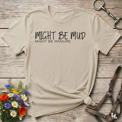 A beige graphic t-shirt with the text 'MIGHT BE MUD / MIGHT BE MANURE' printed in black, styled in a flat-lay on a rustic wooden background with a horse bit, horse halter, and wildflowers.