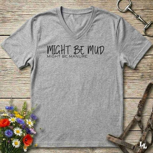 A heather gray V-neck t-shirt with black printed text reading 'MIGHT BE MUD' and 'MIGHT BE MANURE.' laid flat on a rustic wood surface with flowers and horse tack props around it.