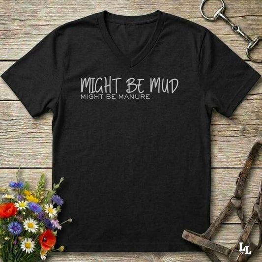 A black V-neck t-shirt with white text reading 'MIGHT BE MUD' and 'MIGHT BE MANURE' on the chest, laid flat on a rustic wooden surface with equestrian-themed props and flowers.