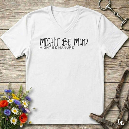 A white v-neck t-shirt with black printed text reading 'MIGHT BE MUD' and 'MIGHT BE MANURE.' laid flat on a rustic wood background with decorative flowers and horse tack around it.