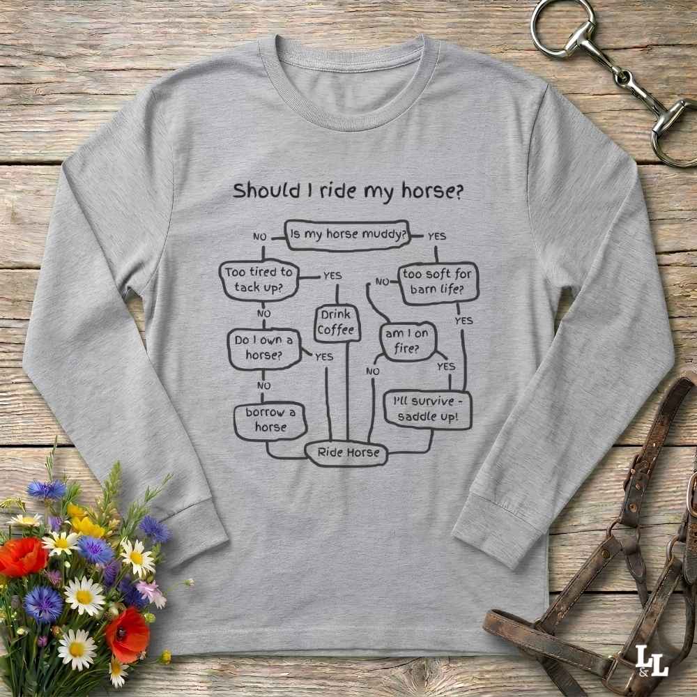A light gray long-sleeve t-shirt with a black flowchart graphic reading 'Should I ride my horse?' styled in a flat-lay on a rustic wooden background with a bouquet of flowers and leather tack.