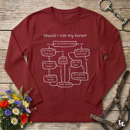 A long-sleeve maroon t-shirt with a white printed flowchart graphic titled 'Should I ride my horse?' laid flat on a rustic wooden surface. The shirt is styled with equestrian accessories (a leather bridle) and a small bouquet of wildflowers.