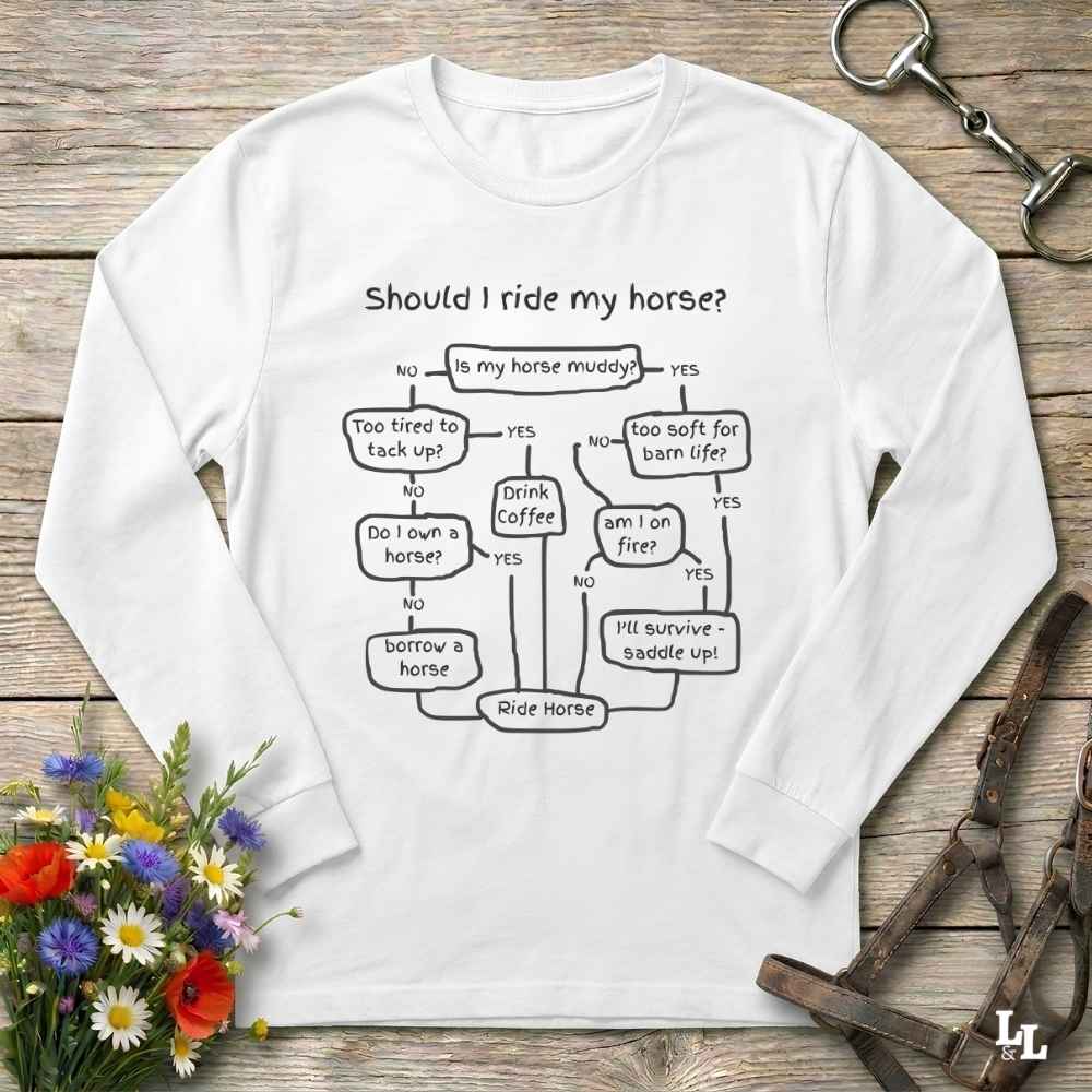 A white long-sleeve crewneck sweatshirt with a black printed flowchart graphic titled 'Should I ride my horse?'. The sweatshirt is displayed in a flat-lay arrangement on a rustic wooden background, accompanied by a bouquet of colorful wildflowers, a leather bridle, and a horse bit.