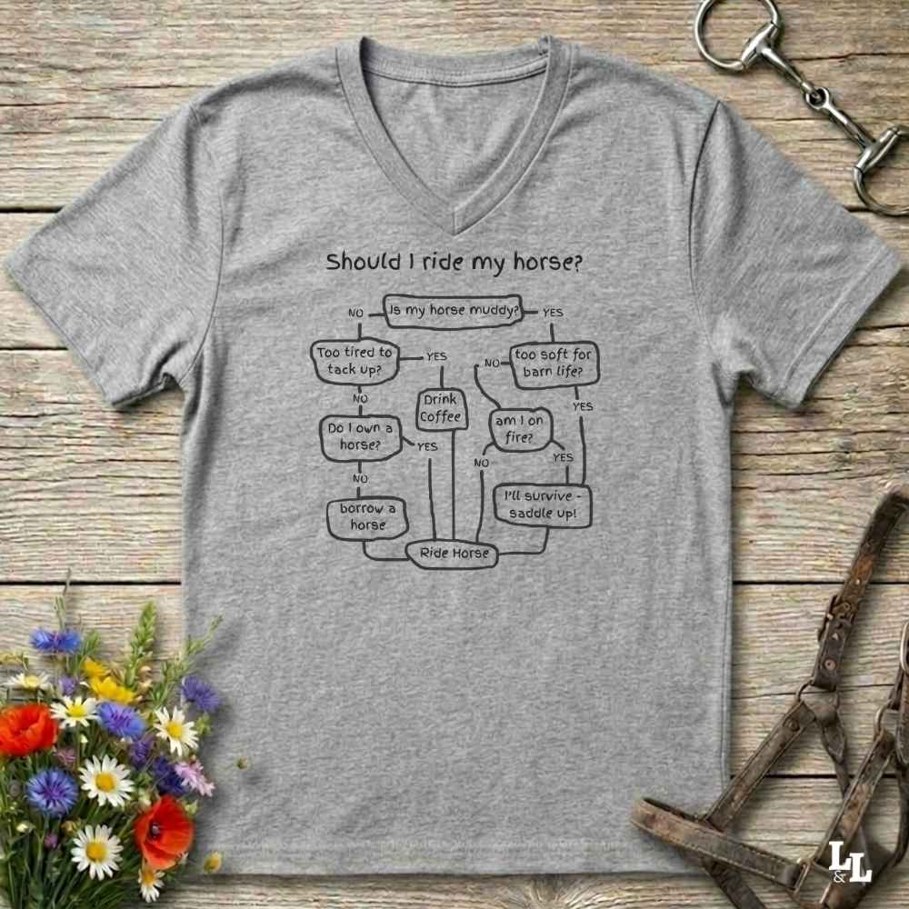 A heather gray V-neck t-shirt with a black printed flowchart titled 'Should I ride my horse?' laid flat on a rustic wooden surface. The shirt is styled with equestrian props including a leather bridle, horse bits, and a small bouquet of wildflowers.
