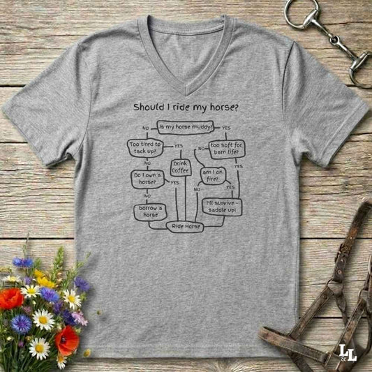 A heather gray V-neck t-shirt with a black printed flowchart titled 'Should I ride my horse?' laid flat on a rustic wooden surface. The shirt is styled with equestrian props including a leather bridle, horse bits, and a small bouquet of wildflowers.