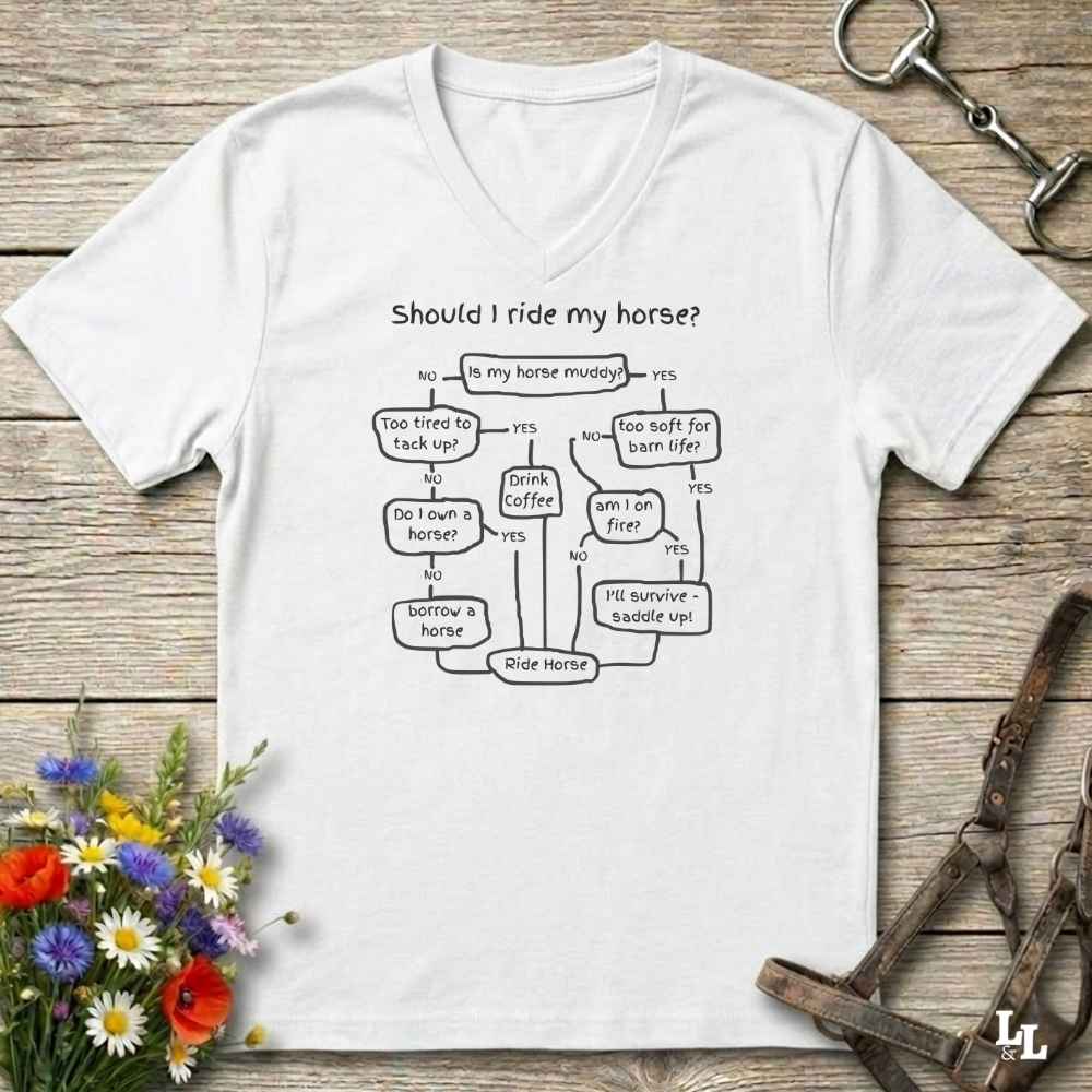 A white V-neck t-shirt with a black printed flowchart graphic titled 'Should I ride my horse?' laid flat on a rustic wooden surface. The shirt is styled with a horse bit, a small bouquet of wildflowers, and a metal horse tack strap in the corners.