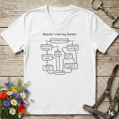 A white V-neck t-shirt with a black printed flowchart graphic titled 'Should I ride my horse?' laid flat on a rustic wooden surface. The shirt is styled with a horse bit, a small bouquet of wildflowers, and a metal horse tack strap in the corners.