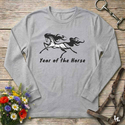 A light gray long-sleeve t-shirt with a black graphic of a rearing horse and the text 'Year of the Horse' printed on the front, displayed on a rustic wooden background with decorative equestrian and floral props.