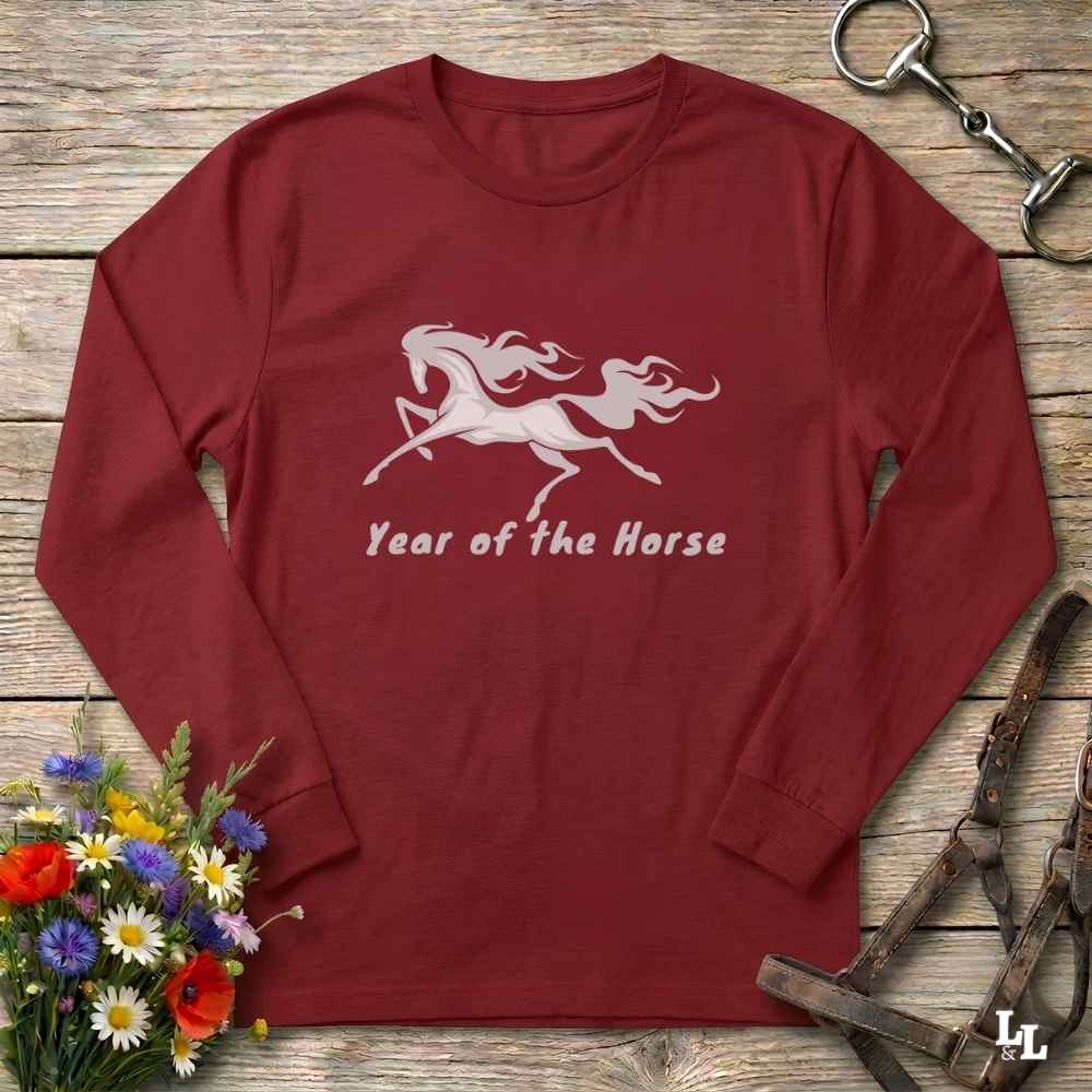 A long-sleeve crewneck t-shirt in maroon red, featuring a white graphic of a rearing horse and the text 'Year of the Horse' printed on the chest. The shirt is displayed flat on a rustic wooden background, styled with equestrian leather tack, a metal bit, and a small bouquet of colorful wildflowers.