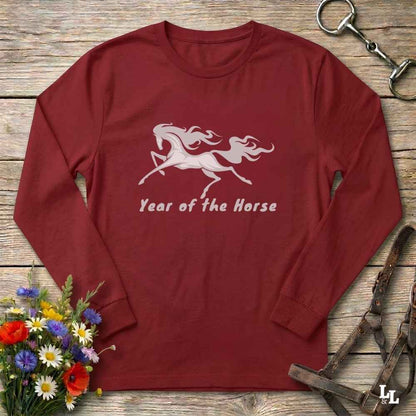 A long-sleeve crewneck t-shirt in maroon red, featuring a white graphic of a rearing horse and the text 'Year of the Horse' printed on the chest. The shirt is displayed flat on a rustic wooden background, styled with equestrian leather tack, a metal bit, and a small bouquet of colorful wildflowers.