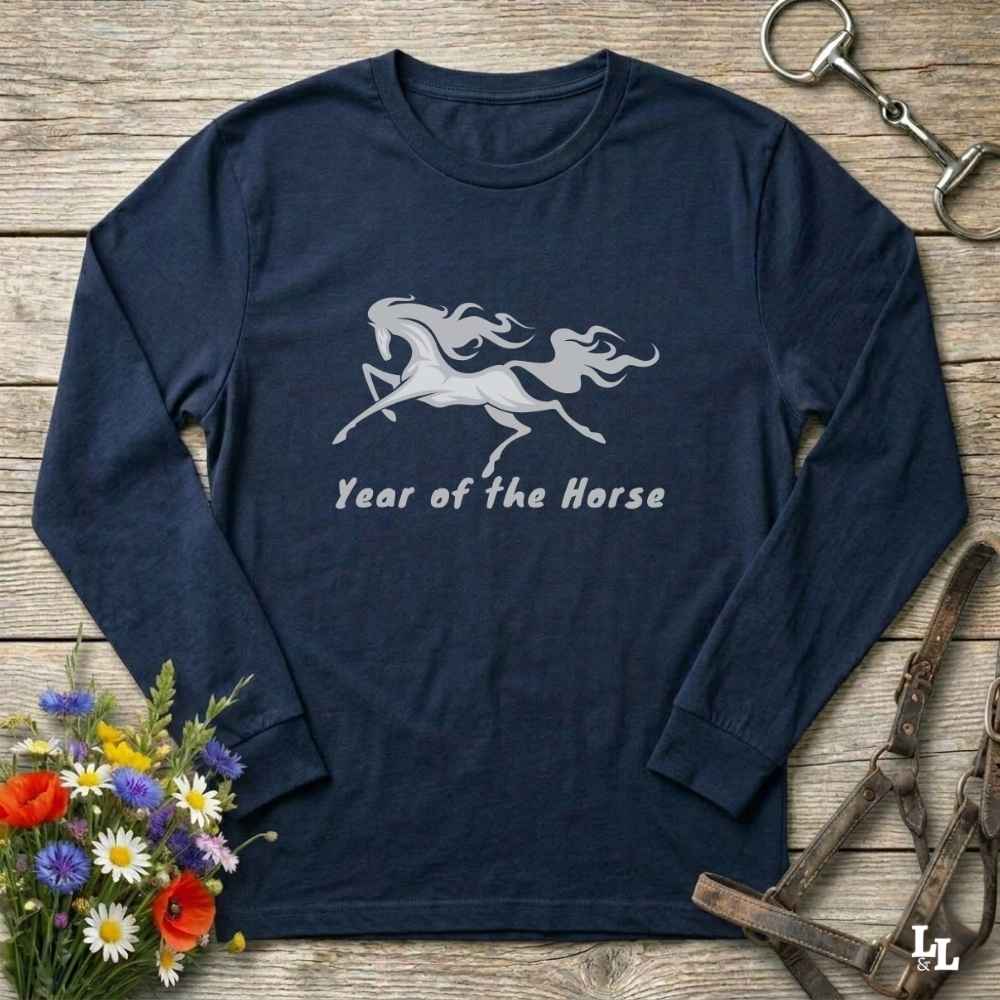 A navy blue long-sleeve t-shirt with a white graphic of a running horse and the text 'Year of the Horse' printed on the chest. The shirt is displayed in a flat-lay arrangement on a rustic wooden background, surrounded by equestrian-themed props including a horse bit, a halter, and a small bouquet of colorful wildflowers.