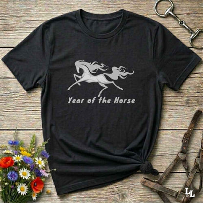 A black short-sleeve t-shirt with a white graphic of a running horse and the text 'Year of the Horse' printed on the front, styled flat on a rustic wooden background with equestrian accessories and flowers.