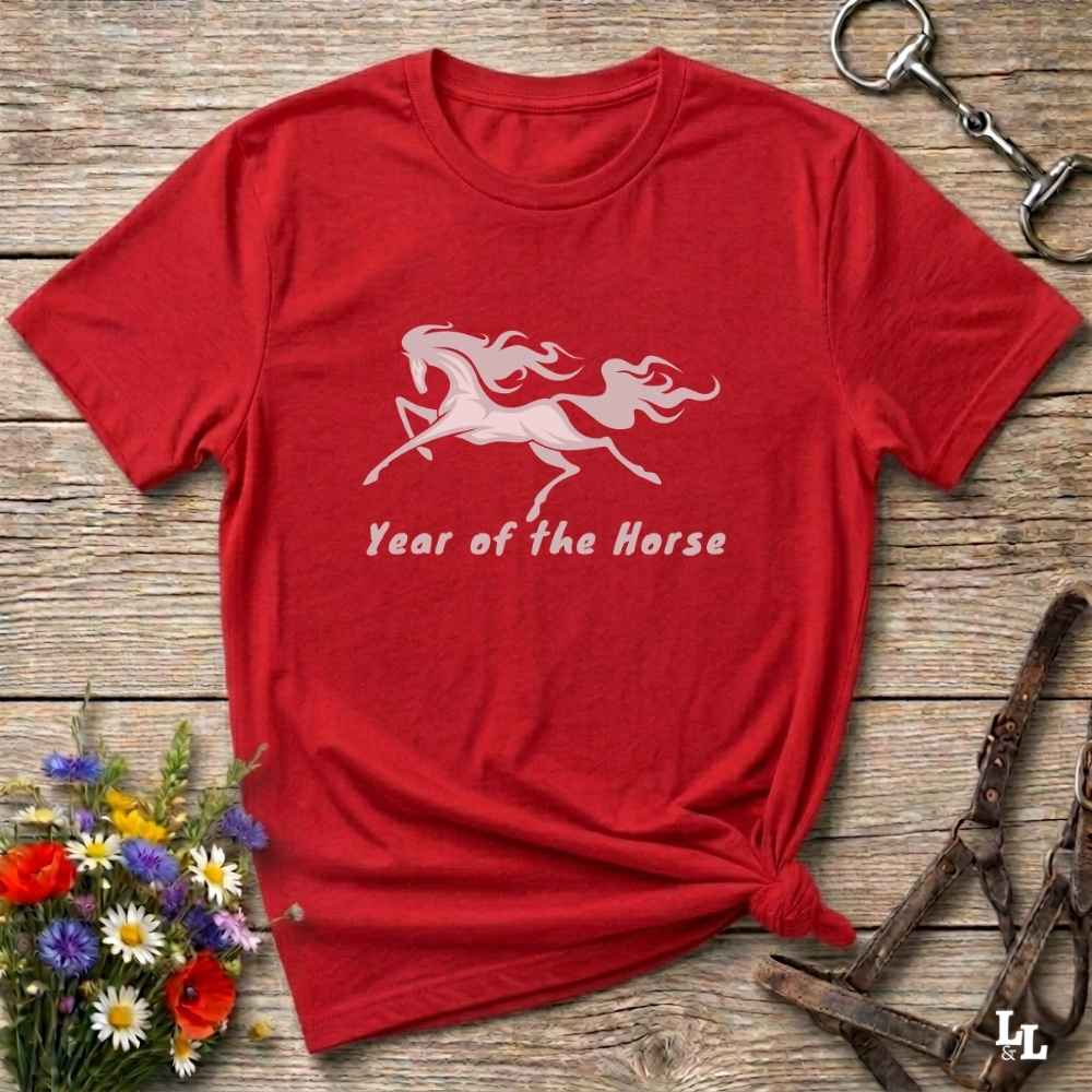 A red short-sleeve t-shirt with a white graphic of a rearing horse and the text 'Year of the Horse' printed on the front, styled flat on a rustic wooden background with equestrian accessories and flowers.