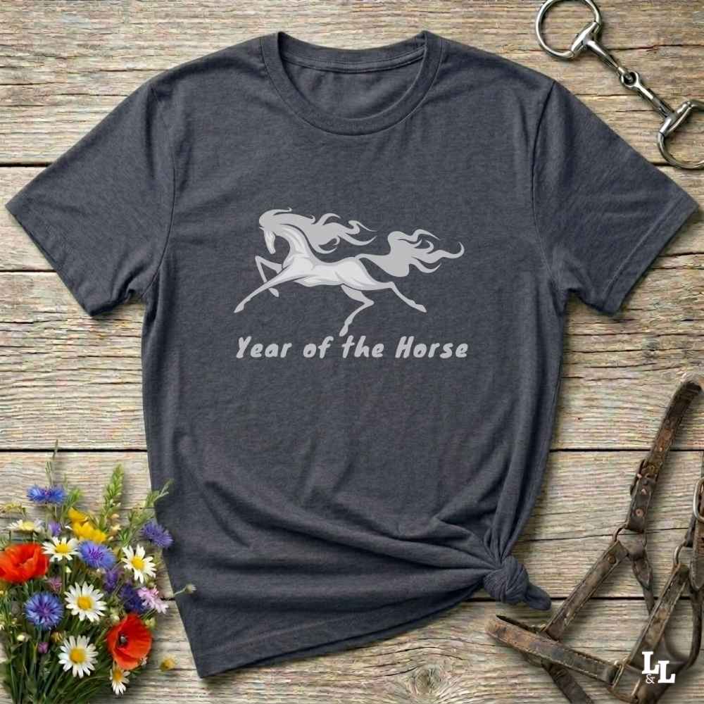 A dark heather gray t-shirt with a white graphic of a rearing horse and the text 'Year of the Horse' printed on the front. The shirt is styled in a flat-lay arrangement on a rustic wooden background, surrounded by equestrian-themed accessories (a halter, a horse bit, and a small bouquet of wildflowers).