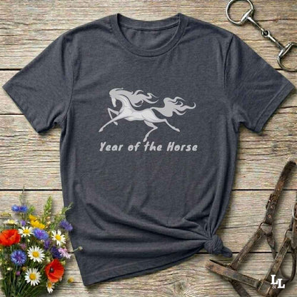 A dark heather gray t-shirt with a white graphic of a rearing horse and the text 'Year of the Horse' printed on the front. The shirt is styled in a flat-lay arrangement on a rustic wooden background, surrounded by equestrian-themed accessories (a halter, a horse bit, and a small bouquet of wildflowers).