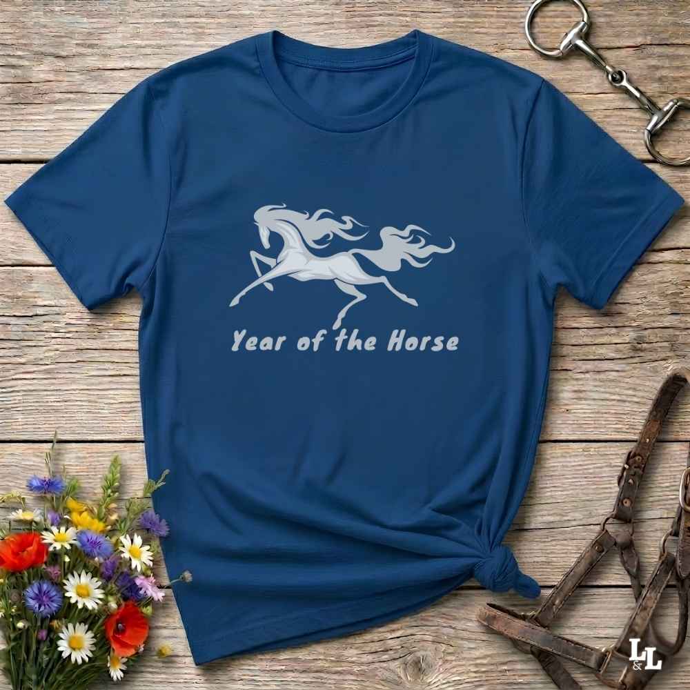A navy blue short-sleeve t-shirt with a white graphic of a rearing horse and the text 'Year of the Horse' printed on the front. The shirt is styled in a flat-lay arrangement on a rustic wooden background, accompanied by a small bouquet of colorful flowers, a brown leather halter, and a metal keyring.