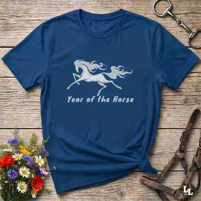 A navy blue short-sleeve t-shirt with a white graphic of a rearing horse and the text 'Year of the Horse' printed on the front. The shirt is styled in a flat-lay arrangement on a rustic wooden background, accompanied by a small bouquet of colorful flowers, a brown leather halter, and a metal keyring.