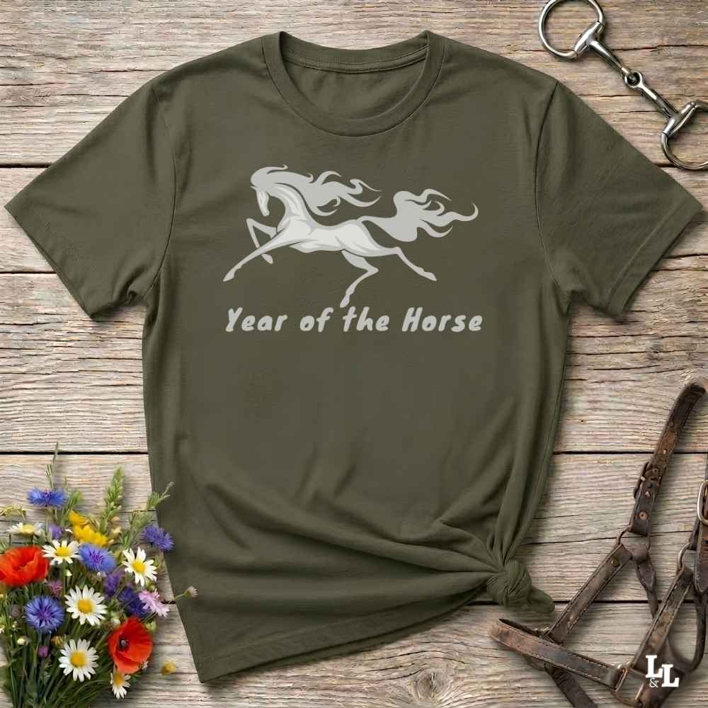 An olive green short-sleeve t-shirt with a white graphic of a galloping horse and the text 'Year of the Horse' printed on the front. The shirt is styled in a flat-lay arrangement on a rustic wooden background, surrounded by decorative elements including a small bouquet of wildflowers, a horse bit, and a leather halter.