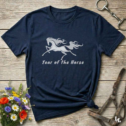 A navy blue short-sleeve t-shirt with a white graphic of a rearing horse and the text 'Year of the Horse' printed on the front. The shirt is styled in a flat-lay arrangement on a rustic wooden background, surrounded by equestrian-themed props including a halter, tack, and a small bouquet of wildflowers.
