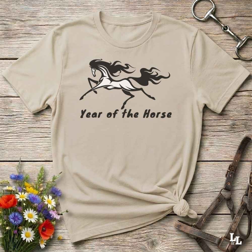 A beige graphic t-shirt with a black horse silhouette and the text 'Year of the Horse' printed on the front, styled in a flat-lay on a rustic wooden background with equestrian accessories and flowers.