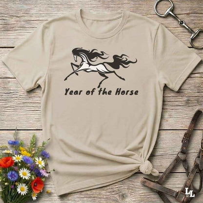 A beige graphic t-shirt with a black horse silhouette and the text 'Year of the Horse' printed on the front, styled in a flat-lay on a rustic wooden background with equestrian accessories and flowers.