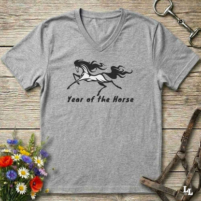 A heather gray V-neck t-shirt with a black graphic of a running horse and the text 'Year of the Horse' printed on the front. The shirt is displayed flat on a rustic wooden background with equestrian-themed props like a horse bit, leather bridle, and a small bouquet of wildflowers.