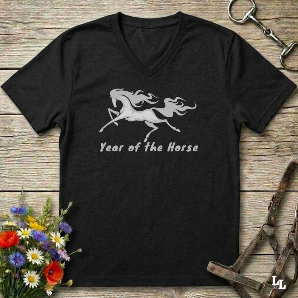 A black V-neck t-shirt with a gray graphic of a rearing horse and the text 'Year of the Horse' printed on the chest, displayed flat on a rustic wooden background with equestrian accessories and flowers.