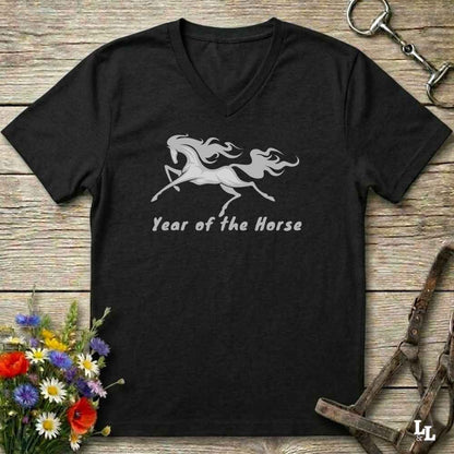 A black V-neck t-shirt with a gray graphic of a rearing horse and the text 'Year of the Horse' printed on the chest, displayed flat on a rustic wooden background with equestrian accessories and flowers.