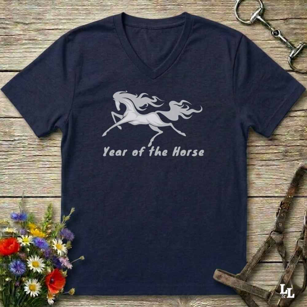 A navy blue V-neck t-shirt with a white graphic of a rearing horse and the text 'Year of the Horse' printed on the chest, displayed flat on a rustic wooden background with equestrian accessories and flowers.