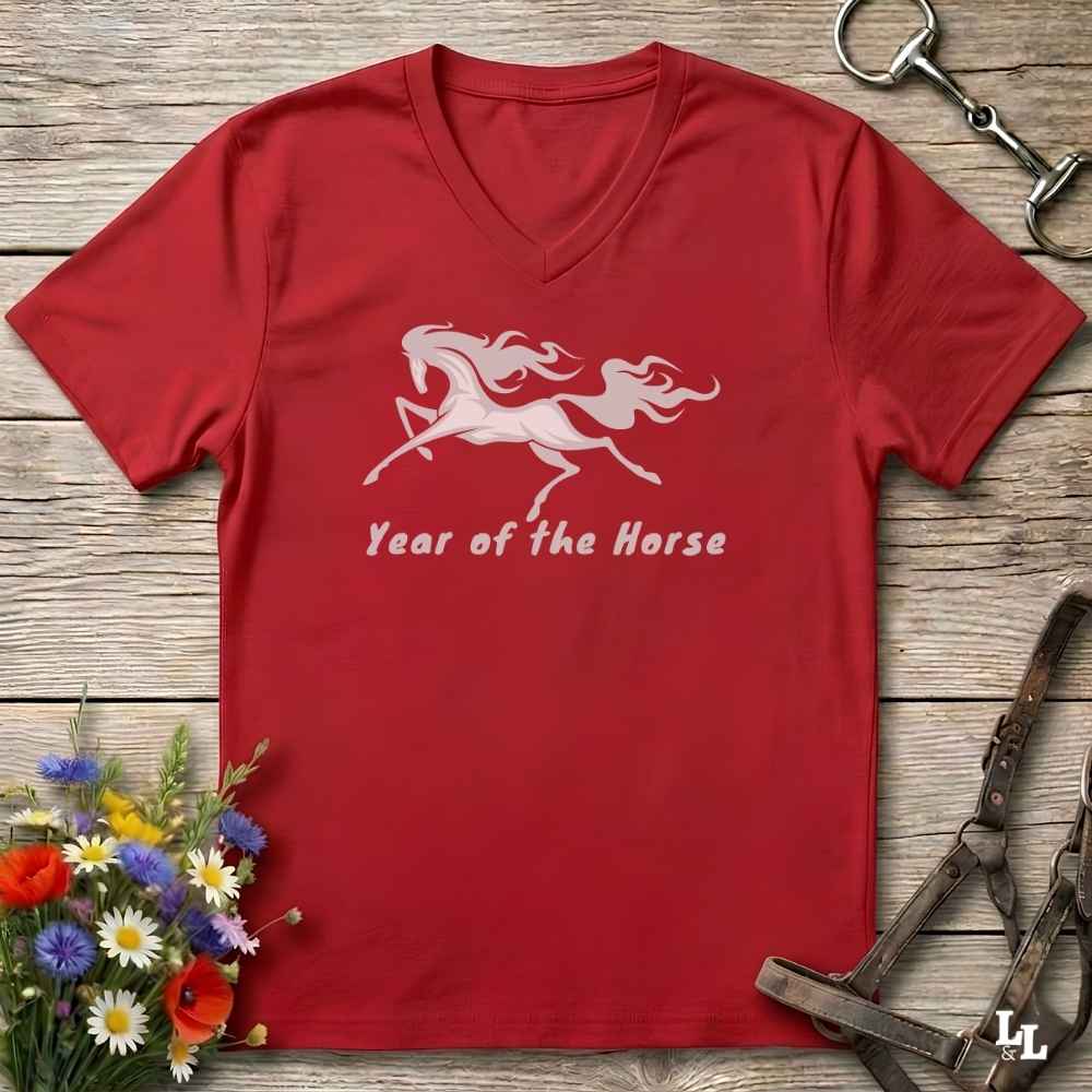A red V-neck t-shirt laid flat on a rustic wooden surface, featuring a white graphic of two leaping horses and the text 'Year of the Horse'. The shirt is styled with equestrian tack, a small bouquet of colorful wildflowers, and metal horse bits.